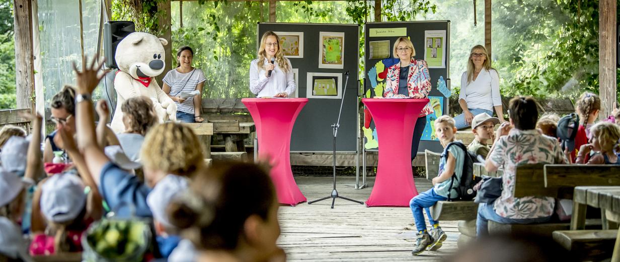 Eine Kindergruppe sitzt auf Bänken. Im Hintergrund stehen zwei Personen an Stehtischen und halten Mikrofone in der Hand.