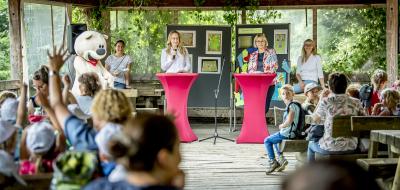 Eine Kindergruppe sitzt auf Bänken. Im Hintergrund stehen zwei Personen an Stehtischen und halten Mikrofone in der Hand.