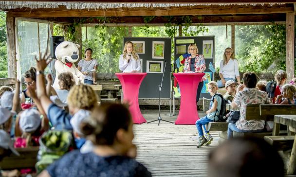 Eine Kindergruppe sitzt auf Bänken. Im Hintergrund stehen zwei Personen an Stehtischen und halten Mikrofone in der Hand.